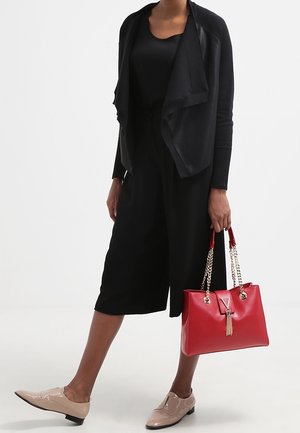 Cardigan noir, combinaison noire, ballerines nudes et un sac à main rouge avec des accents de chaîne. Le sac à main présente un détail en frange.