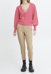 Cárdigan de punto rosa con un escote en V profundo, cinco botones y mangas globo, combinado con pantalones ajustados beige y botas negras con cordones.