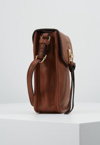 Borsa a tracolla in pelle marrone con una chiusura dorata, rifiniture nere e una tracolla regolabile, poggiata su una superficie bianca.