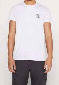 Camiseta blanca de algodón con mangas cortas que presenta un pequeño gráfico de texto manuscrito en el lado izquierdo del pecho. Cuello redondo, diseño sencillo.