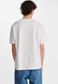 Biała bawełniana koszulka z krótkim rękawem, okrągłym dekoltem, gładką strukturą i prostym wykończeniem. Widoczny szew na plecach i luźny fason.