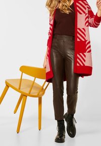 Frau in rotem, gemustertem Cardigan, die einen gelben Holzstuhl hält, braune Lederhose und schwarze Schnürstiefel trägt, geht nach vorn.