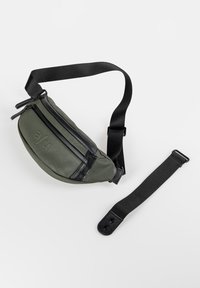 Groene leren heuptas met zwarte accenten, twee met ritsen afgesloten vakken en een verstelbare band, met een gladde textuur en een gebogen vorm.