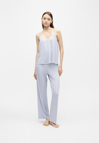 Jeune femme debout pieds nus portant un caraco bleu clair et un pantalon de pyjama ample sur fond blanc.