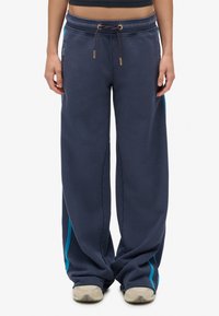 Persoon in losse donkerblauwe sweatpants met turquoise zijkanten en beige sneakers, staande tegen een witte achtergrond.