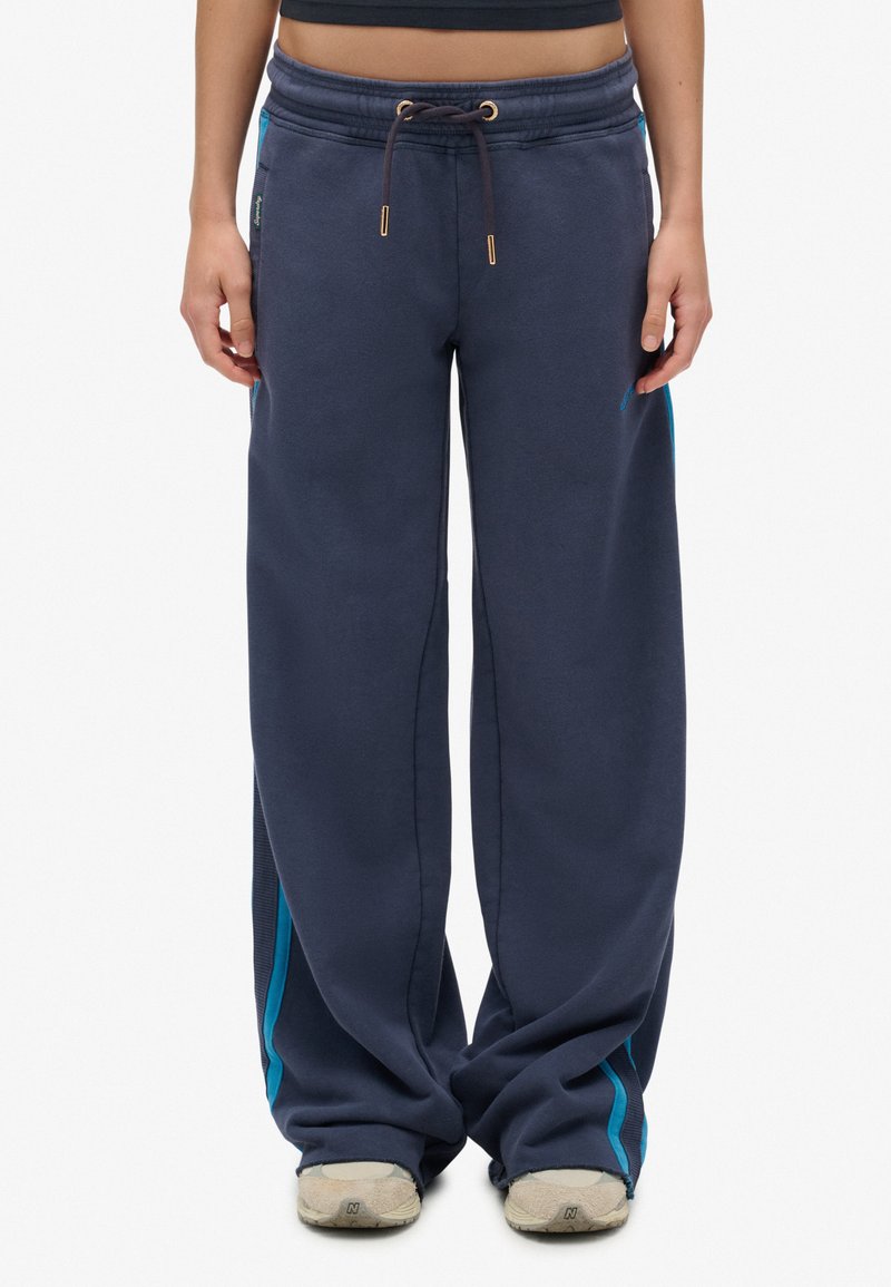 Persoon in losse donkerblauwe sweatpants met turquoise zijkanten en beige sneakers, staande tegen een witte achtergrond.