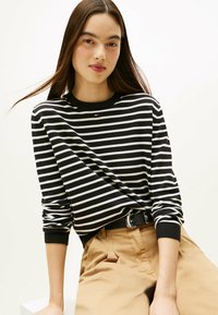 Schwarz-weiß gestreiftes Langarmshirt mit rundem Halsausschnitt, getragen zu khakifarbenen Hosen und einem schwarzen Gürtel. Glatte Stoffstruktur.