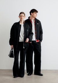 Γυναίκα και άνδρας φορούν μαύρα μπουφάν και παντελόνια· η γυναίκα κρατάει μαύρη τσάντα χειρός· ο άνδρας φοράει κόκκινο καρό πουκάμισο κάτω από το μπουφάν, και οι δύο στέκονται σε απλό φόντο.