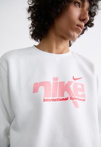 Sweatshirt branco com um grande logótipo "Nike" em vermelho pontilhado e o texto "International Sportswear". Material suave de algodão com decote redondo.