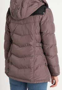 Giacca isolante viola con cappuccio, dotata di un pannello superiore nero, design trapuntato, tasche laterali e vita regolabile.