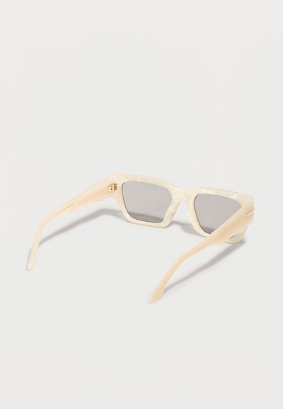 Γυαλιά ηλίου σε σχήμα γατίσιου ματιού με εκρού χρωματισμό και γκρι φακούς, απεικονιζόμενα από οπίσθια τριχοτομημένη γωνία σε απλό λευκό φόντο.