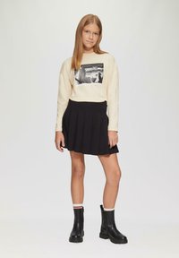 Langarm-Cremeshirt mit einem schwarz-weißen Grafikdesign, kombiniert mit einem schwarzen Faltenrock und schwarzen Stiefeletten mit strukturiierter Sohle.