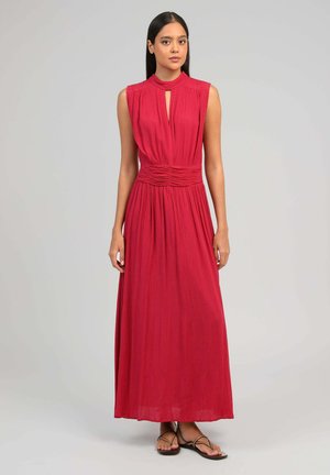Femme debout portant une robe maxi rouge sans manches en tissu plissé, taille cintrée, encolure goutte d'eau et sandales plates noires à brides.