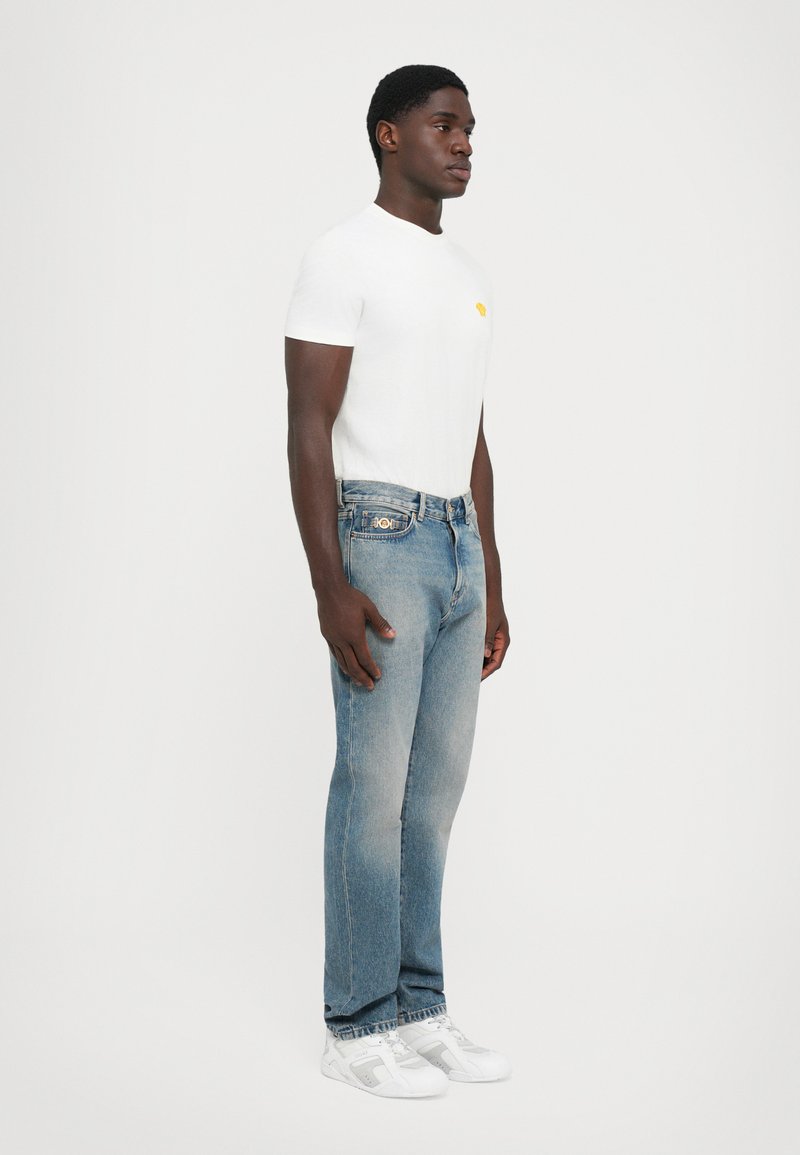 Man som står i vit t-shirt, blå jeans med ett guldlogotyp på fickan och vita sneakers mot en enkel ljus bakgrund.
