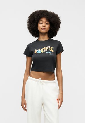 Γυναίκα με σγουρά μαλλιά που φοράει μαύρο κομμένο t-shirt με την επιγραφή «Pacific Coast» και λευκό παντελόνι με κορδόνι, στέκεται μπροστά σε απλό φόντο.