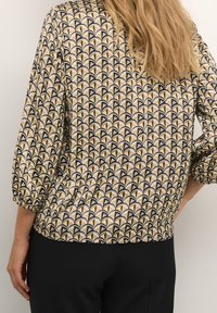 Mujer vestida con una blusa de patrón de abanico en beige y negro con mangas de tres cuartos y pantalones negros, de pie contra una pared clara y lisa.