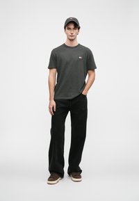 Mannlig modell iført mørkegrå t-skjorte med logo, svarte vide jeans, svart caps, og brune joggesko, stående mot en enkel lys grå bakgrunn.