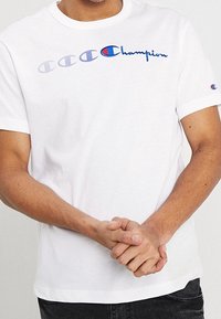 White cotton t-shirt featuring a blue "Champion" logo and repeated logo design, short sleeves, and crew neckline.