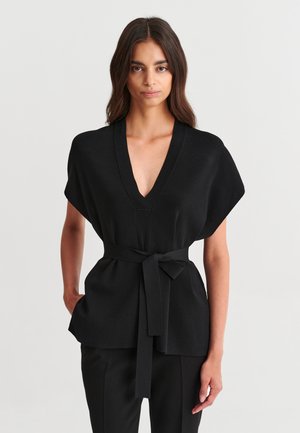 Femme aux longs cheveux foncés portant un haut noir à col en V avec de larges manches et une ceinture à la taille, associé à un pantalon noir, debout devant un fond uni.