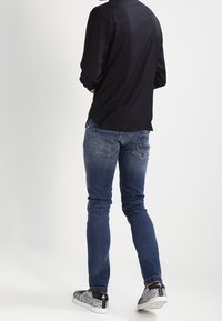 Uomo che indossa una camicia scura a maniche lunghe, jeans blu e sneakers a motivo bianco e nero, in piedi con le spalle rivolte verso lo spettatore su uno sfondo bianco.