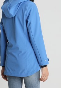 Persona con una chaqueta azul ajustada con capucha y jeans celestes, vista desde atrás con los brazos relajados a los lados.