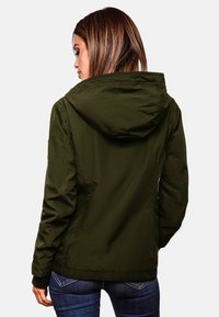 Groene waterdichte jas met capuchon, gemaakt van een textuurstof, met een getailleerde pasvorm en elastische manchetten, gedragen over denim jeans.
