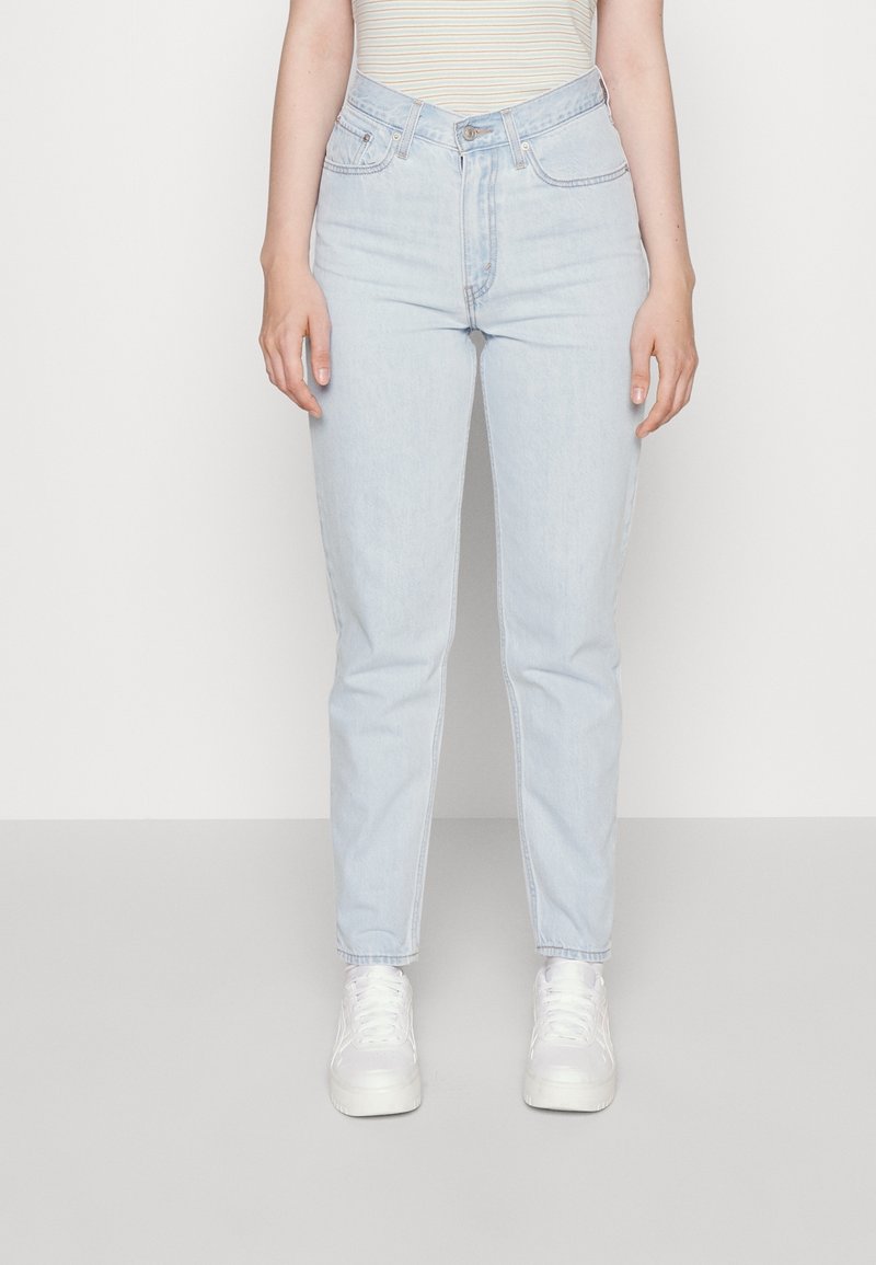 Femme portant un jean bleu clair taille haute à jambes droites, des baskets blanches et une chemise beige rayée sur un fond uni clair.