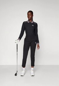 Svart golfoutfit med en topp med krage och rosa detaljer, tajta byxor och en logotyp. Personen håller i en golfklubba och bär vita skor.
