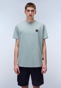 T-shirt in cotone verde chiaro con maniche corte, scollatura rotonda e piccolo logo circolare sul petto. Indossato con pantaloni corti blu navy.