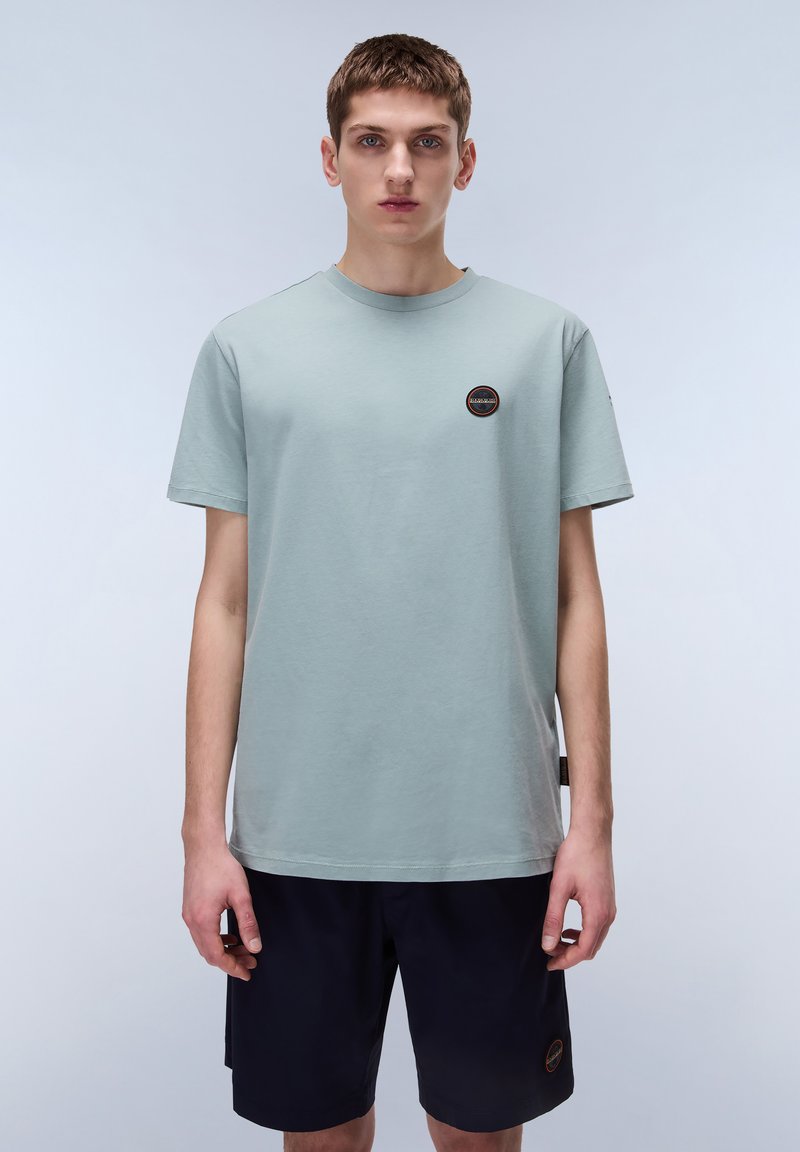 T-shirt in cotone verde chiaro con maniche corte, scollatura rotonda e piccolo logo circolare sul petto. Indossato con pantaloni corti blu navy.