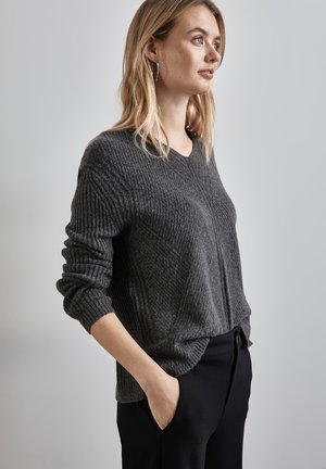 Pull en maille côtelée gris avec un col en V, coupe décontractée et manches longues ; porté sur un pantalon noir, mettant en avant la texture et le design décontracté.
