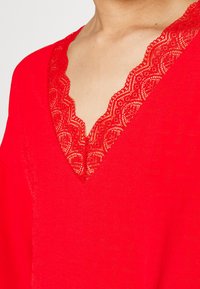 Rotes Oberteil mit tiefem V-Ausschnitt und aufwändiger Spitzenverzierung, aus einem weichen Material gefertigt, das eine glatte Textur und ein leichtes Design präsentiert.