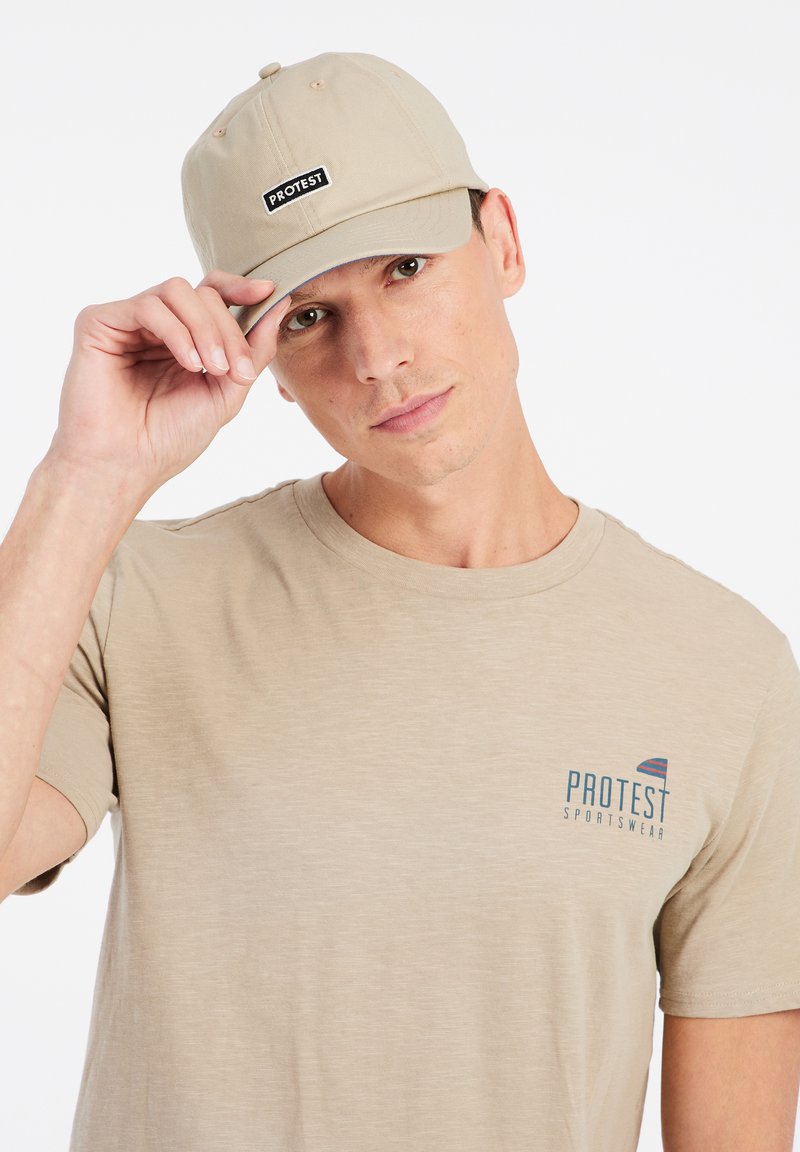 Khaki baseballcap met een gestikt zwart "PROTEST" logo. Het model draagt een lichtbeige T-shirt met een klein meerkleurig logo.