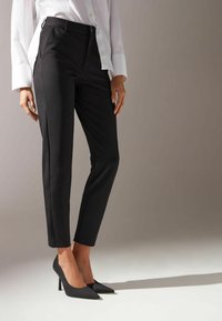 Pantalones negros ajustados con un corte slim y textura suave, con una cintura de altura media, pliegue frontal sutil y bolsillos laterales. Combinados con tacones negros.