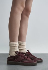 Burgundy sneakers with suede and leather textures, featuring beige ribbed socks. Low-top design with lace closure and contrasting accents.