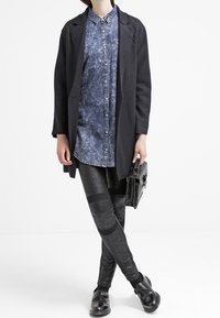 Cappotto lungo nero sopra una camicia di denim, abbinato a leggings neri texturizzati con dettagli sulle ginocchia e scarpe nere, mentre tiene una borsa nera.