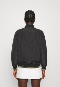 Personne aux cheveux tressés et aux boucles d'oreilles en anneau porte une veste bomber noire et une jupe blanche, debout, vue de dos sur un fond blanc.