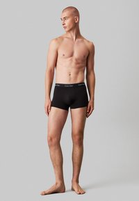 Zwarte katoenen boxershorts met een brede elastische tailleband waarop het "Calvin Klein" logo staat. Model staat blote voeten, met een neutrale grijze achtergrond.