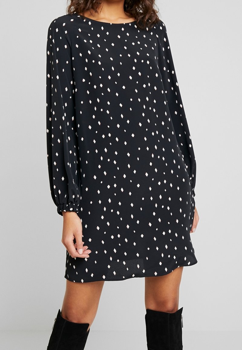 Femme portant une mini-robe noire à manches longues avec des motifs de diamants blancs et des bottes noires montantes jusqu’aux genoux, debout devant un fond uni.