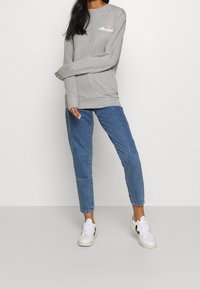 Femme portant un sweat-shirt gris ellesse, un jean en denim bleu et des baskets blanches, debout contre un fond clair uni, les bras croisés.