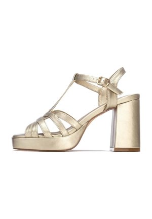 Sandalia de tacón alto metálica dorada con tacón cuadrado, con múltiples tiras entrecruzadas y hebilla ajustable en el tobillo. Textura suave.