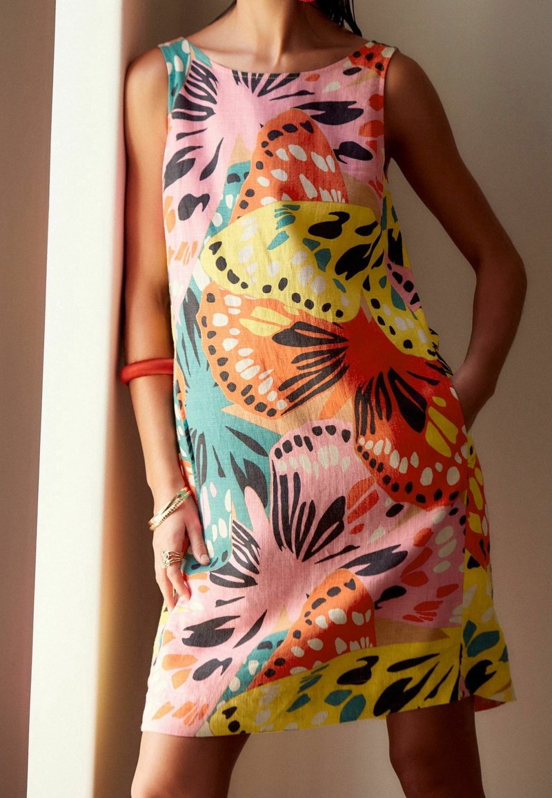 Robe sans manches aux motifs de papillons colorés et abstraits, portée par une personne debout, une main dans la poche et des bijoux sur l'autre bras.