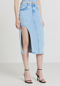 Jupe en denim bleu clair avec une fente devant, ourlet brut et design classique à cinq poches. Associée à des sandales à talons hauts noires à lanières.
