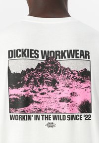 Camiseta de algodón blanca con un gráfico en negro y rosa de un paisaje rocoso, texto "DICKIES WORKWEAR" y "WORKIN' IN THE WILD SINCE '22."