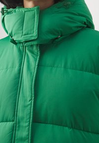 Gepolsterte grüne Jacke mit hohem Kragen, ausgestattet mit einer glatten Textur, Reißverschluss und genähten Details auf der Brust.