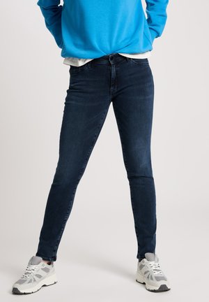 Dunkelblaue Skinny Jeans mit glatter Textur, mittelhoher Taille und subtilen Nähten, kombiniert mit einem hellblauen Sweatshirt und Sneakers.