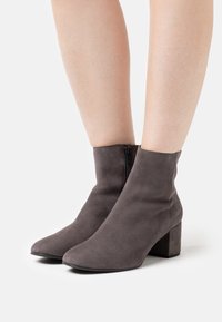 Šedé semišové kotníkové boty s špičatou špičkou, bočním zipem a silným podpatkem. Design je elegantní a těsně obepíná kotník.