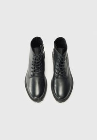 Botas de tobillo de cuero negro con cordones y cremalleras laterales, mostradas desde arriba sobre un fondo blanco. Acabado suave y puntera redondeada.
