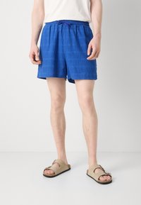 Pantalones cortos azules de textura con un patrón a cuadros, que cuentan con una cintura elástica y cordón. Llevados con sandalias beige de tipo slip-on.