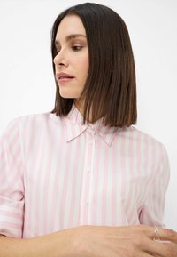 Light pink striped blouse with short sleeves, button closure, collar, and smooth texture. Notable white buttons contrast with the fabric.